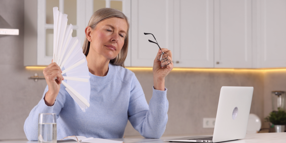 A middle-aged woman sits at a desk with a laptop and a glass of water, holding a hand fan and her glasses while experiencing a hot flash.
